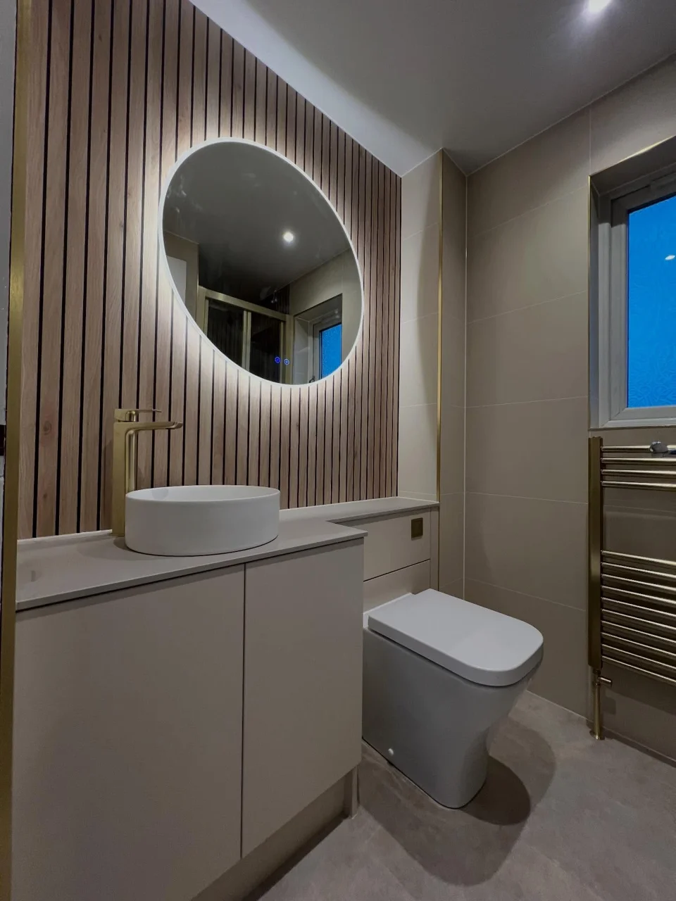 Round illuminated mirror above a white basin and brass tap with wood-effect wall panelling
