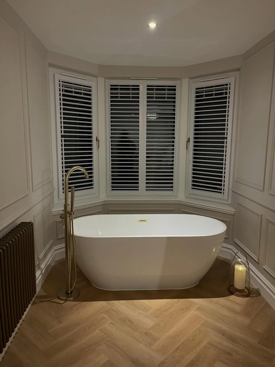 White freestanding bath below bay-window shutters with a brass floor-standing tap