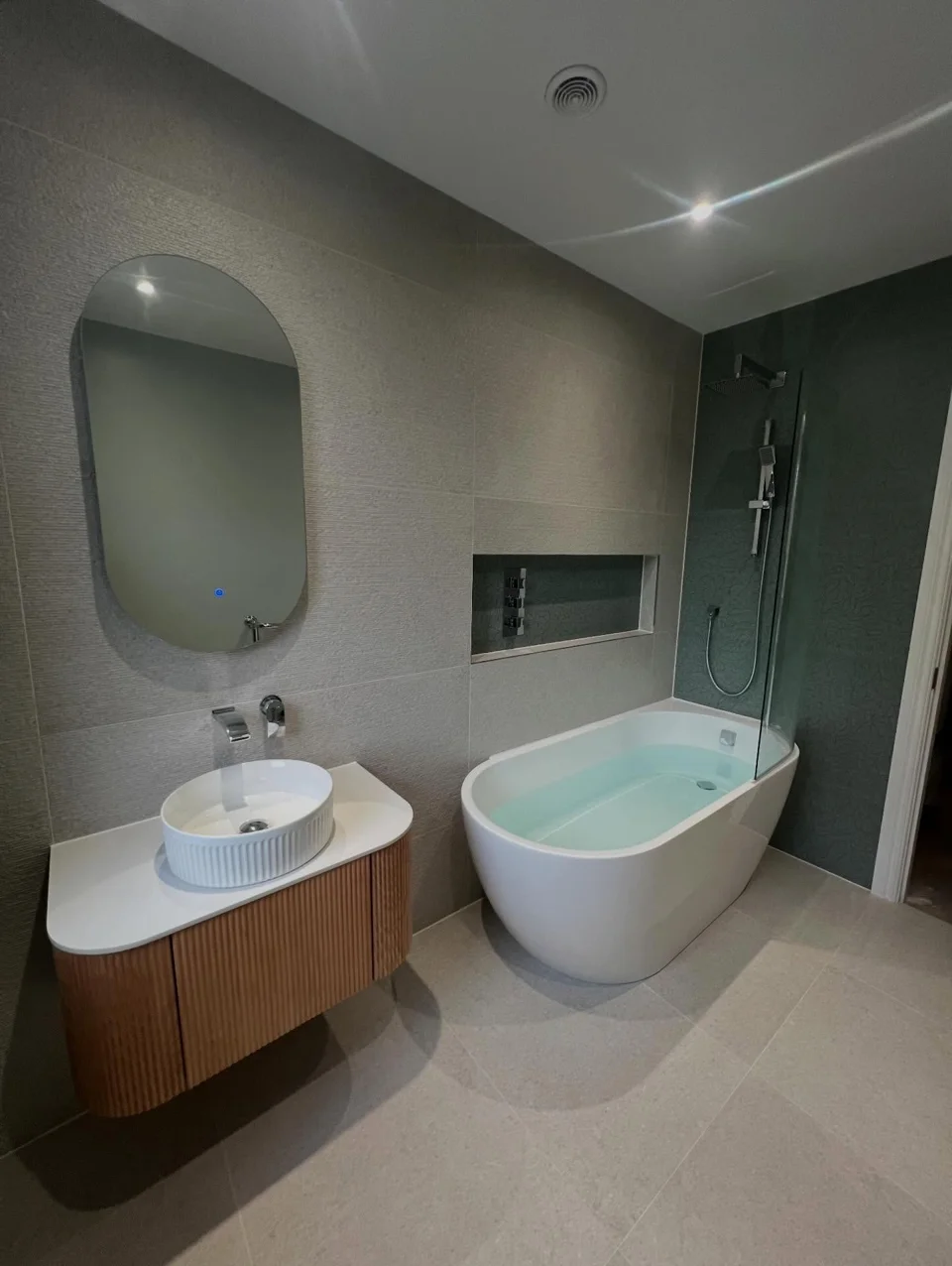 Freestanding bath beside a wall-mounted timber vanity, with a shower screen and recessed controls
